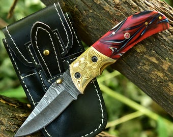 May include: A folding knife with a patterned blade and a red, black, and white handle. The handle has gold accents and is paired with a black leather sheath with white stitching. The knife is resting on a tree branch.