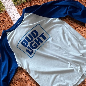 Könnte beinhalten: Ein weißes und blaues Baseball-Shirt mit den Worten "BUD LIGHT" in einem blauen Rechteck. Die Ärmel sind einfarbig blau.