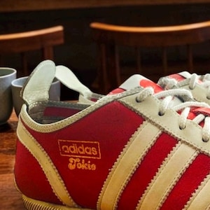 Op de afbeelding: Rode en crèmekleurige Adidas Tokio sneakers. De sneakers hebben het Adidas-logo en het woord "Tokio" in gouden letters. Ze hebben witte veters en een crèmekleurige zool. De achtergrond is wazig en toont een houten tafel en stoelen.