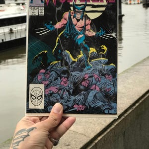Puede incluir: Una portada de cómic con Wolverine, un personaje de Marvel Comics, de pie sobre una pila de soldados. La portada tiene el título "Wolverine" en letras grandes de color naranja. El cómic lo sostiene una persona con un tatuaje.