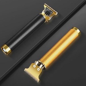 Op de afbeelding: Twee elektrische haartrimmers met T-vormige bladen. De ene trimmer heeft een zwarte, getextureerde cilindrische handgreep met gouden accenten, terwijl de andere een volledig gouden, getextureerde handgreep heeft. Beide apparaten hebben metalen snijkoppen.
