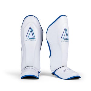 May include: Pair of white shin guards with blue trim and the "ANTARES" logo in blue. The shin guards are designed for athletic use and have a curved shape for a comfortable fit. The text "ANTARES ATHLETICS" is printed on the bottom.
