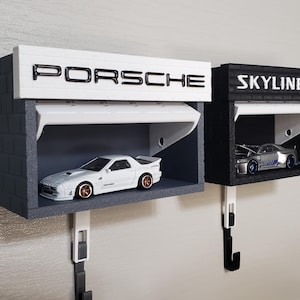 Puede incluir: Dos estantes de exhibición de coches en miniatura, uno con la etiqueta "PORSCHE" y el otro "SKYLINE GT-R". Cada estante tiene un coche en miniatura en su interior. El estante Porsche es blanco y gris, y el estante Skyline es negro y gris.