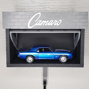 Op de afbeelding: Een blauwe modelauto wordt tentoongesteld in een zwarte, garage-achtige structuur. Het woord "Camaro" staat in witte letters boven de auto. De garage heeft een grijze bakstenen gevel.