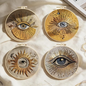 May include: Four circular embroidery hoops with celestial designs. Each features an eye or sun and moon motif, with gold and silver thread detailing. The designs are set against a neutral fabric background, creating a textured, handcrafted appearance.