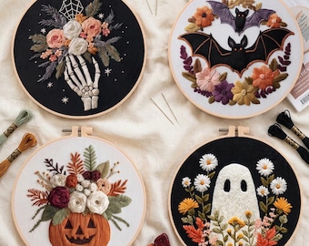 May include: Four circular embroidery hoops featuring seasonal designs: a skeleton hand holding flowers on black fabric, two bats surrounded by autumn flowers on white, a jack-o'-lantern with floral accents, and a white ghost among daisies on black.