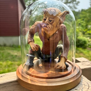 May include: A detailed sculpture of a goblin-like creature inside a glass dome. The figure is brown with pointed ears and a furrowed brow, wearing a brown shirt and green pants. The dome sits on a wooden base, creating a unique display piece.