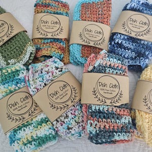 May include: A collection of handmade dish cloths in various colors, including green, blue, yellow, and multi-colored blends. Each cloth is wrapped with a brown paper band that reads "Dish Cloth 100% Pure Cotton Handmade". The cloths are made of a knitted or crocheted material.