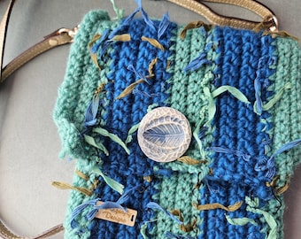 Può includere: Una borsa all'uncinetto fatta a mano nei toni del blu e del verde acqua, ornata da frange decorative. La borsa presenta un bottone rotondo trasparente con un disegno a foglia e una tracolla dorata. Una piccola etichetta recita "Dragonfly Designs."
