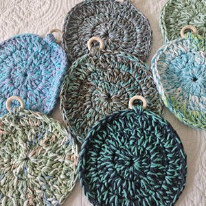 May include: A collection of handmade, circular crochet dishcloths in various colors, including blue, green, and gray. Each cloth features a wooden ring for hanging. The crochet work is visible, showcasing the texture and craftsmanship of the items.