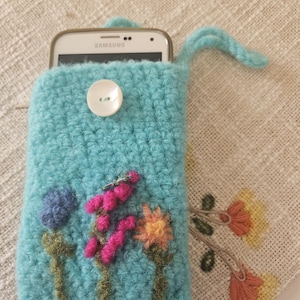 May include: A turquoise crocheted phone case with a white button closure and a loop handle. The case features embroidered flowers in pink, orange, and blue. A white smartphone is partially visible inside the case.