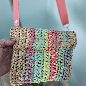 May include: A woven crossbody bag with a flap closure. The bag features a tan base with colorful vertical stripes in pink, blue, yellow, and green. It has a pink strap and a small tag that reads "Dragonfly Designs".