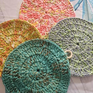 May include: Five circular, handmade crochet dishcloths in a variety of colours. The dishcloths feature a textured stitch pattern. Colours include pink, yellow, green, and teal. One dishcloth has a wooden ring attached. Perfect for kitchen use.