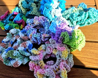 May include: A collection of colorful, handmade crochet scrunchies in various shades of blue, green, pink, purple, and yellow. The scrunchies are arranged on a wooden surface, showcasing their textured, looped design. These hair accessories are perfect for adding a pop of color.