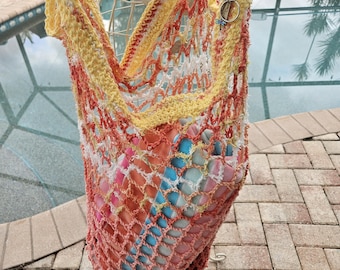 May include: A handmade, open-weave crochet tote bag in shades of yellow, orange, and red. The bag is filled with colorful items and has a dragonfly charm attached. The bag is displayed on a wire mannequin.