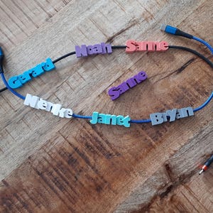 Könnte beinhalten: USB-Ladekabel mit personalisierten Namensschildern. Die Kabel sind schwarz und blau, mit Namen wie "Gerard", "Noah" und "Janet" in verschiedenen Farben. Auf einer Holzoberfläche angeordnet.