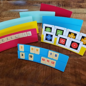 May include: A collection of colorful game pieces, including red, yellow, blue, and teal holders with tiles and cards. The tiles display letters and numbers, while the cards feature geometric shapes. The word "GEZELIG" is spelled out on one of the holders.
