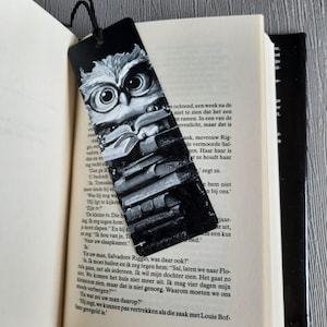 Puede incluir: Marcapáginas en blanco y negro con un búho con gafas sobre una pila de libros. El marcapáginas está insertado en un libro con texto visible. Una cuerda negra está unida a la parte superior.