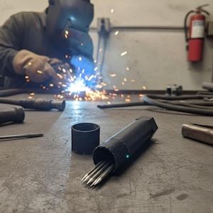 Puede incluir: Una herramienta de metal negro con múltiples puntas, junto a una pieza cilíndrica a juego, descansa sobre una superficie metálica. Una persona soldando en el fondo crea chispas. Otras herramientas están dispersas.
