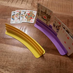 May include: Two curved playing card holders, one yellow and one purple, with playing cards in them. A deck of cards is next to the yellow holder. The holders have slots to hold the cards upright. The background is a wooden surface.