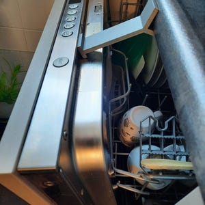 May include: A silver-colored dishwasher door propped open with a light gray plastic bracket. The control panel features a start button and various settings. Inside the dishwasher, a white mug with a black graphic pattern sits on a metal rack.