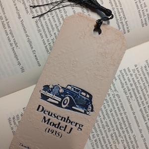 May include: A vintage-style bookmark with a black tassel. The bookmark features a blue illustration of a Duesenberg Model J (1935) car, with the text "Deusenberg Model J (1935)" below the car image. The bookmark is a light beige color.