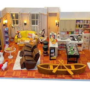 Może przedstawiać: Miniaturowa diorama salonu i kuchni, z żółtą sofą, brązowym fotelem rozkładanym i piłkarzykami. Scena obejmuje małą łódź, białą statuetkę i miniaturową kuchnię ze sprzętem. Ściany są pomalowane na biało, a podłoga jest czerwonobrązowa.