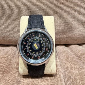 Könnte beinhalten: Eine runde Armbanduhr mit schwarzem Zifferblatt und silberfarbenem Metallgehäuse. Die Uhr hat ein schwarzes Stoffarmband. Das Zifferblatt der Uhr zeigt weiße Ziffern und den Text "PRESENT TIME" und "UNDONE".