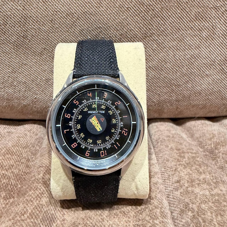 May include: A round wristwatch with a black face and a silver-colored metal case. The watch has a black fabric band. The watch face features white numerals and the text "PRESENT TIME" and "UNDONE".