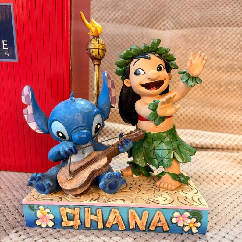 May include: A figurine featuring a blue alien character playing a brown ukulele and a human character wearing a green leaf skirt and lei, standing on a base with the word OHANA and floral accents, next to a tall tiki torch with a yellow flame.