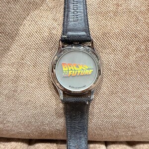 May include: A stainless steel watch with a black leather band. The watch face features the "Back to the Future" logo in yellow and green. The watch is water resistant to 50 meters.