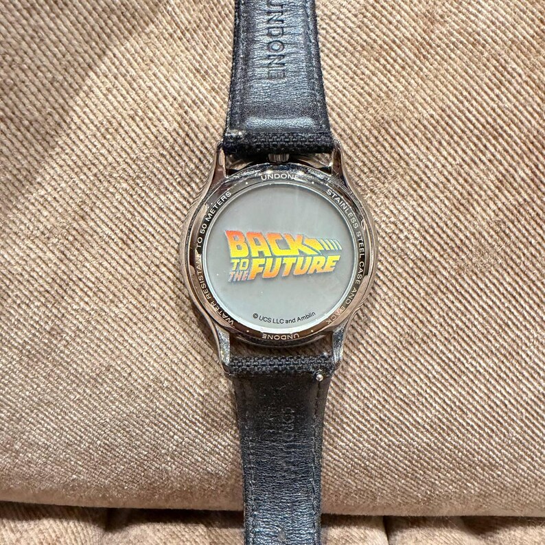 May include: A stainless steel watch with a black leather band. The watch face features the "Back to the Future" logo in yellow and green. The watch is water resistant to 50 meters.