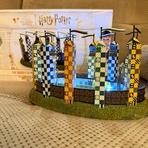 May include: A Harry Potter Quidditch pitch model featuring four tall towers with checkered patterns in yellow, blue, green, and black, topped with small flags. The structure sits on a green base with a dark brown fence and internal LED lighting.