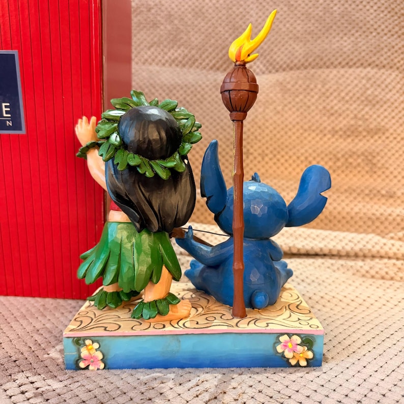May include: A figurine featuring a dark-haired character wearing a green leaf skirt and lei, standing beside a blue creature with large ears. A brown tiki torch with a yellow flame stands between them on a rectangular base with floral accents.