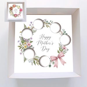 Peut inclure: Une boîte blanche avec un motif de couronne florale et le texte "Happy Mother's Day". La couronne comprend des fleurs roses, violettes et blanches, des feuilles vertes et un nœud rose. Le design comporte six ouvertures circulaires.