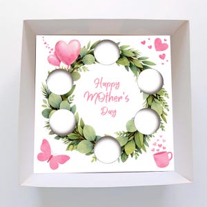 May include: A white box containing a decorative insert for Mother's Day. The insert has a wreath of green leaves and eucalyptus, with six circular cutouts. Pink hearts, a butterfly, and the text "Happy Mother's Day" are included.