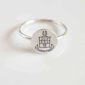 May include: A silver ring with a round, engraved crest featuring a shield with a book, stars, and a bird. The inscription reads "Knowledge is Power".