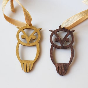 May include: Two wooden owl ornaments with gold ribbon hangers. One is light brown, the other dark brown. The owls have a cutout design with a V-shaped detail on the head and vertical lines at the bottom.