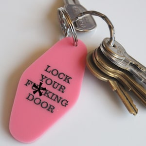 May include: A pink motel-style key tag with the phrase "LOCK YOUR F***ING DOOR" printed in black. A metal keyring holds the tag and several keys of varying sizes and colours.