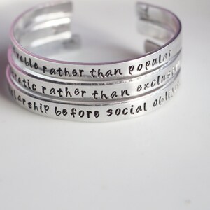 May include: Silver bracelet with four stacked bands, each engraved with a motivational quote: "remarkable rather than popular", "authentic rather than exclusive", "scholarship before social obligation", and "be kind".