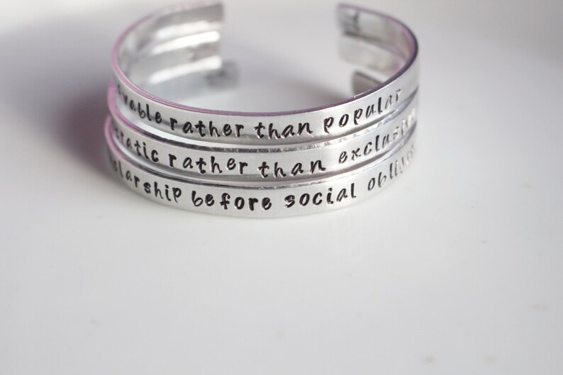 May include: Silver bracelet with four stacked bands, each engraved with a motivational quote: "remarkable rather than popular", "authentic rather than exclusive", "scholarship before social obligation", and "be kind".