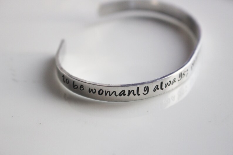May include: Silver bracelet with the inscription "to be womanly always is not to be weak".
