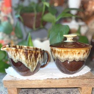 May include: A ceramic creamer pitcher and a lidded sugar bowl featuring a two-tone glaze. The lower sections are a deep, solid brown, while the upper portions transition into a creamy, off-white drip pattern that cascades down the sides.
