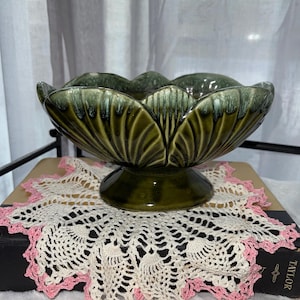 May include: A green ceramic pedestal bowl with a scalloped rim, resting on a white and pink crocheted doily. The bowl has a glossy finish and a leaf-like design. The doily is placed on a dark book.