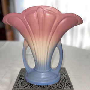 May include: A vintage art deco vase in a stylized floral shape. The vase features a gradient of colors, transitioning from pink at the top to white and then blue at the base. It has two handles and sits on a decorative book.