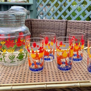 May include: A vintage glass jug and six matching glasses featuring a floral design in red, yellow, and orange. The jug and glasses are on a gold-trimmed rattan tray, ideal for serving drinks. The glasses have a blue base.