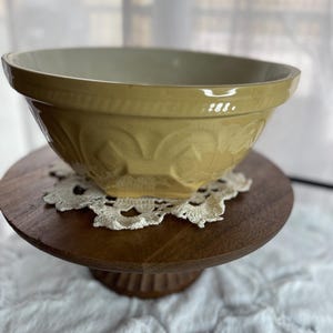 Puede incluir: Un cuenco de cerámica amarilla con diseños florales en relieve, apoyado sobre un soporte de madera para tartas. El cuenco tiene un borde ancho y un interior claro. Un mantelito de encaje blanco se encuentra entre el cuenco y el soporte.