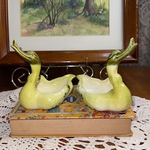 May include: Two ceramic duck figurines with yellow and green coloring. The ducks are perched on a vintage book with a colorful cover. The background includes a framed painting of a landscape scene.
