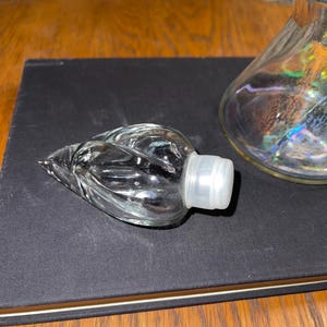 May include: A clear glass bottle stopper with a leaf design and a white plastic base. The stopper is lying on a black surface, with a glass decanter visible in the background. The decanter has an iridescent finish.