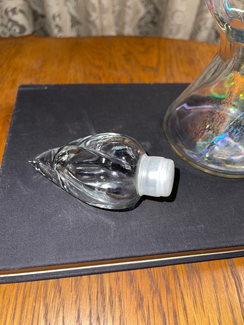 May include: A clear glass bottle stopper with a leaf design and a white plastic base. The stopper is lying on a black surface, with a glass decanter visible in the background. The decanter has an iridescent finish.
